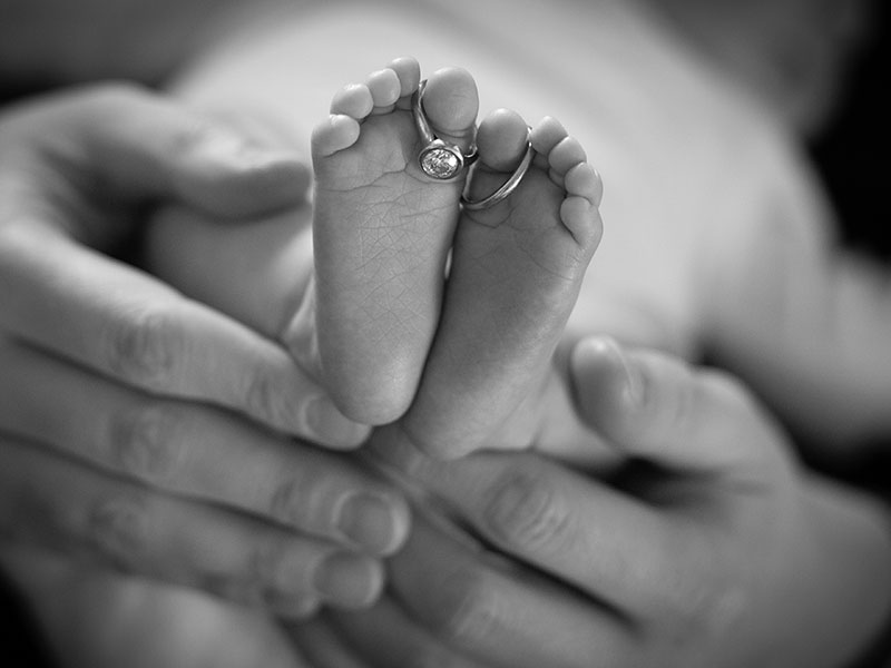 Baby feet is the primary focus, being held by her father's hand. Her parents' wedding rings hang off of her toes.